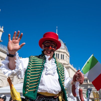 Man wearing traditional Italian dress - red hat, green waistcoat, holding an Italian flag in front of the Exhibition Building.
