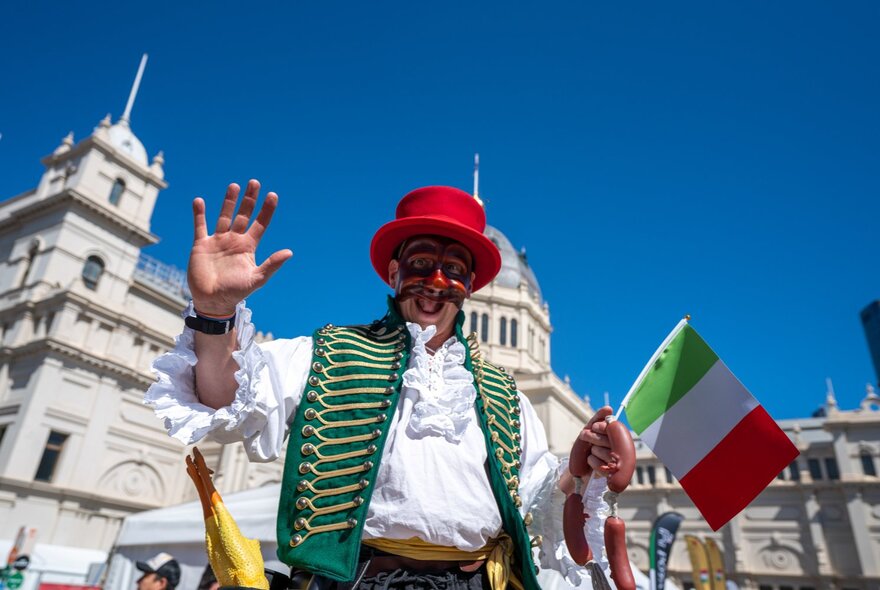 Man wearing traditional Italian dress - red hat, green waistcoat, holding an Italian flag in front of the Exhibition Building.