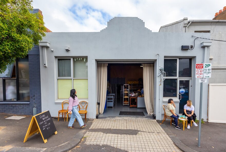 A woman is walking into a bakery from the street. There are two friends sitting on stools out the front eating baked goods.