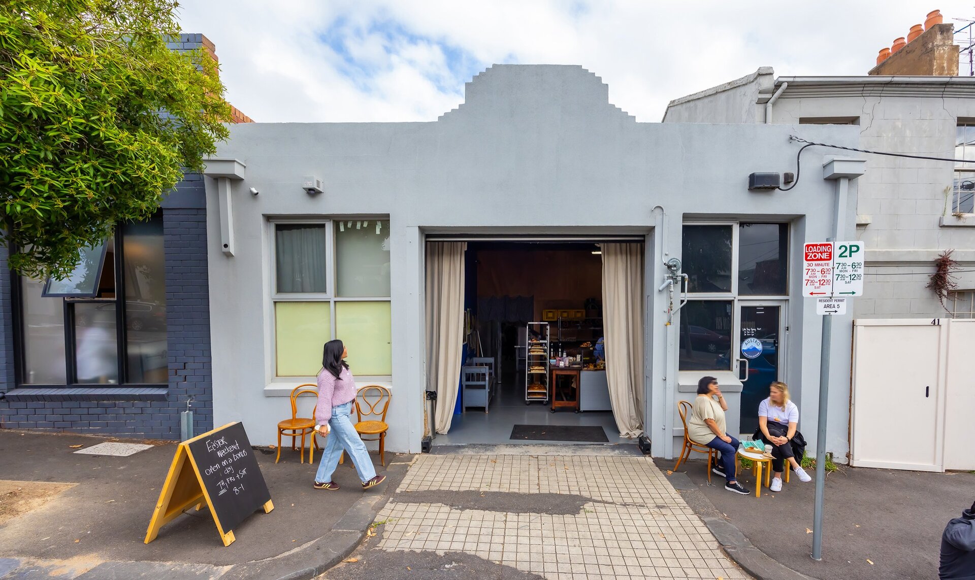 A woman is walking into a bakery from the street. There are two friends sitting on stools out the front eating baked goods.