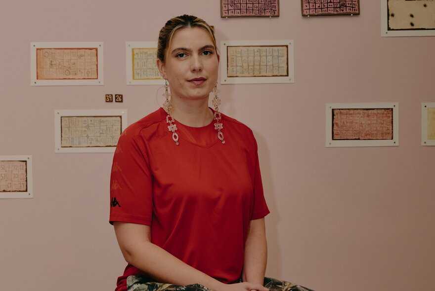 Artist wearing a red T-shirt in a gallery space with small postcard-sized pictures on the wall.