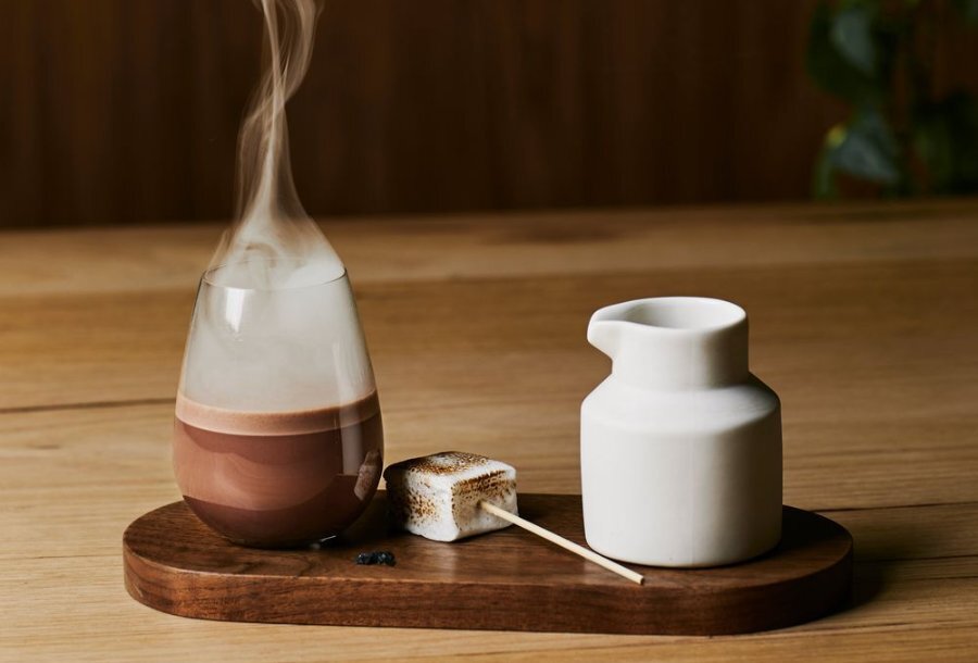 A hot chocolate in a glass with steam coming off it, next to a marshmallow and milk jug. 