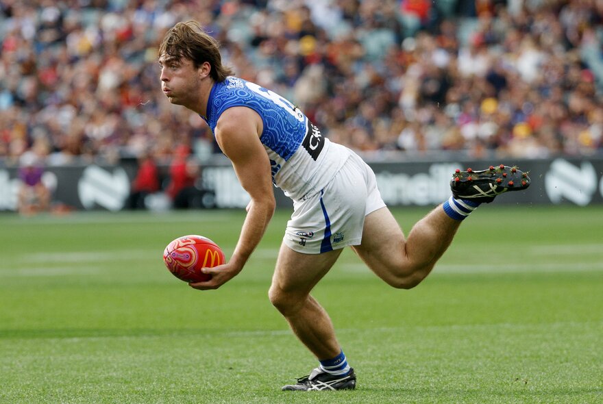 A North Melbourne AFL football player running with the ball during a match, a blurred stadium crowd in the background.