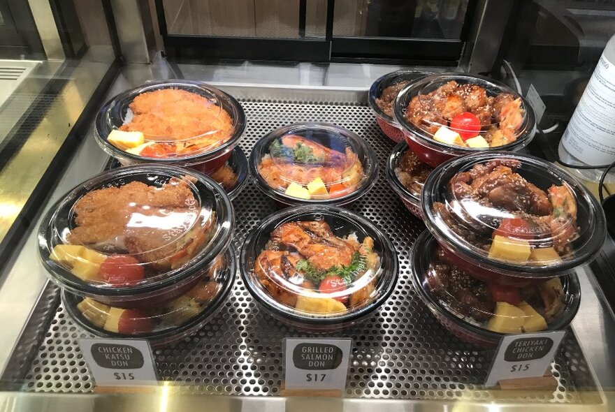 A selection of takeaway Japanese food stacked up in black bowls with clear plastic lids.