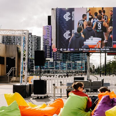 An outdoor event space at Harbour Esplanade Docklands, featuring a large screen broadcasting a sports match to an audience seated in colourful bean bags.