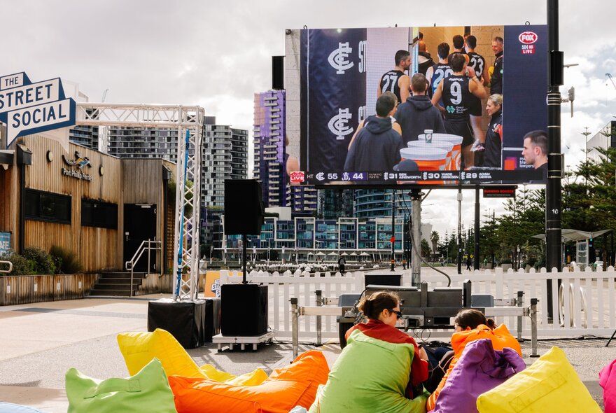 An outdoor event space at Harbour Esplanade Docklands, featuring a large screen broadcasting a sports match to an audience seated in colourful bean bags.