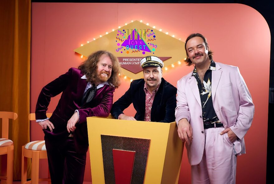 The three hosts of Human Love Quest, all wearing suits, standing behind a pedestal in a set that resembles a TV game show.