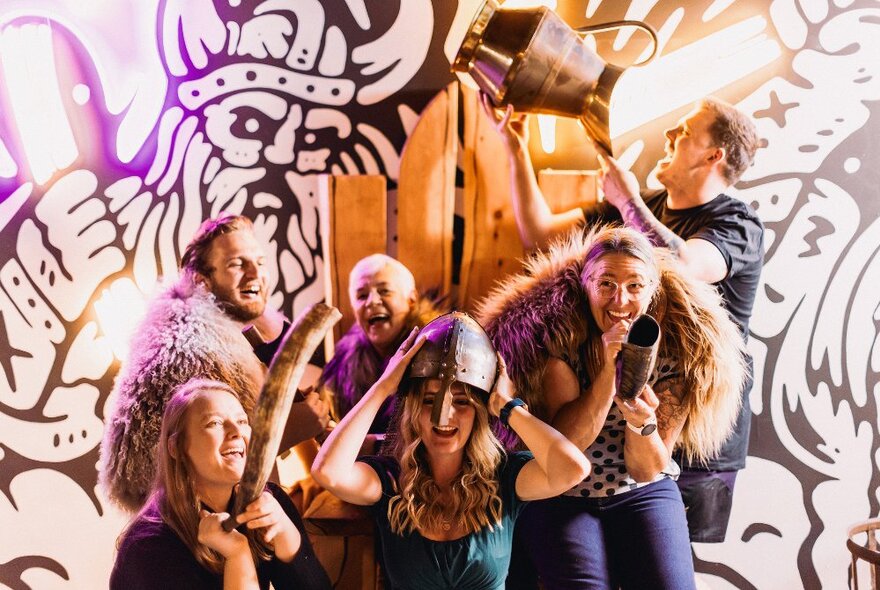 A group of people wearing viking inspired costumes over their normal clothes, laughing, in front of a wall mural of vikings.