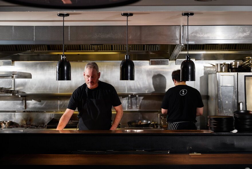 Chef wearing black T-shirt standing behind the pass in a stainless-steel kitchen.