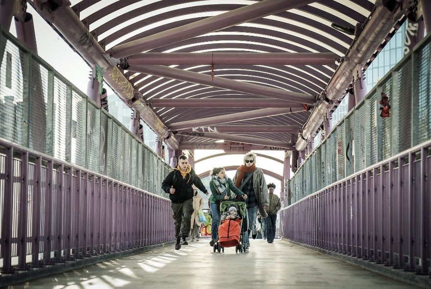 A still image from a film showing people walking through a purplish overpass with a pram. 