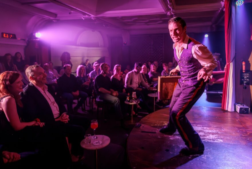 A man wearing a suit on a small stage performing to an audience in a intimate cabaret theatre.