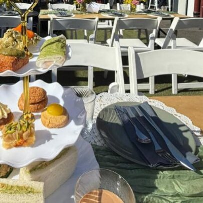 A three tiered plate stand with savoury and sweet small food items on it, at an outdoor table, with tables and chairs in the background.