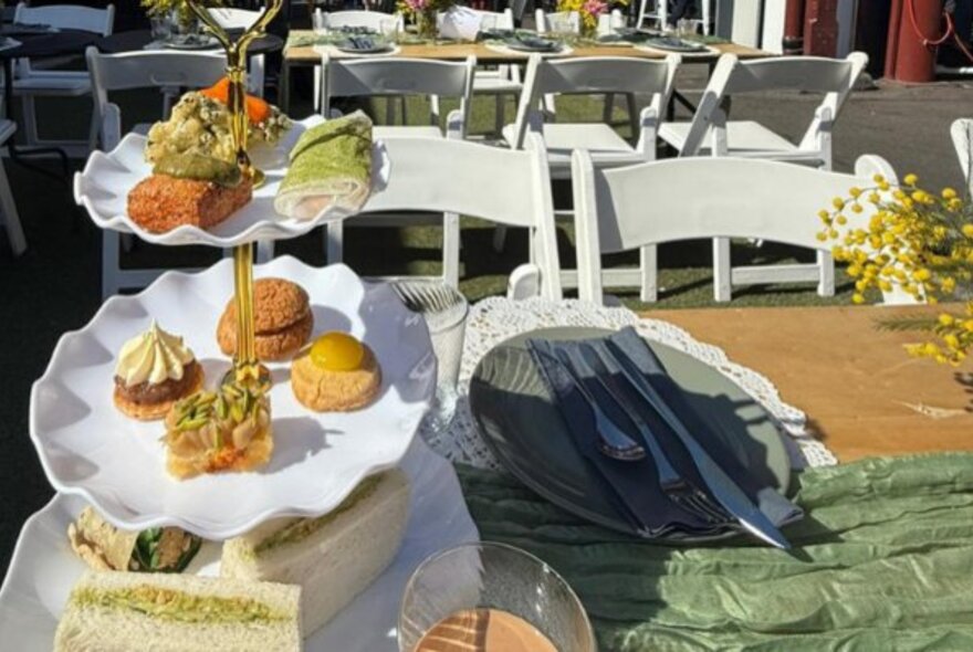 A three tiered plate stand with savoury and sweet small food items on it, at an outdoor table, with tables and chairs in the background.