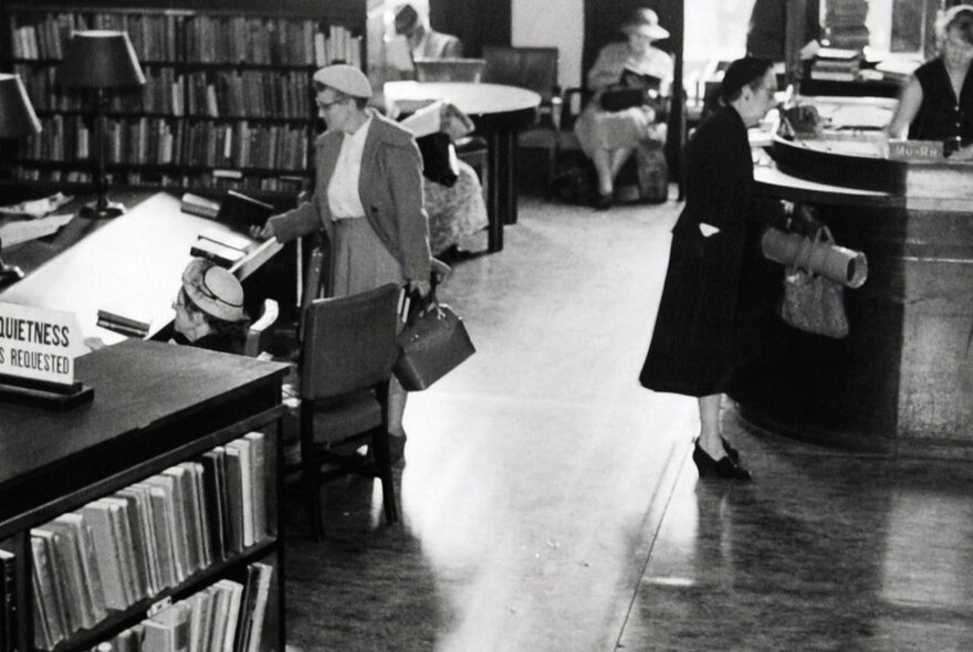 Vintage black and white photograph of Melbourne's Athenaeum Lending Library with people wearing 1940s style clothing seated in chairs or at tables reading, shelves of books arranged in the space.