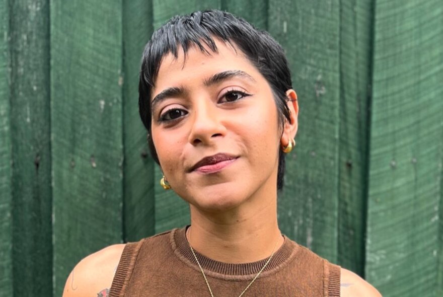 A woman in a brown tank top with a pixie cut smiles in front of a green fence. 