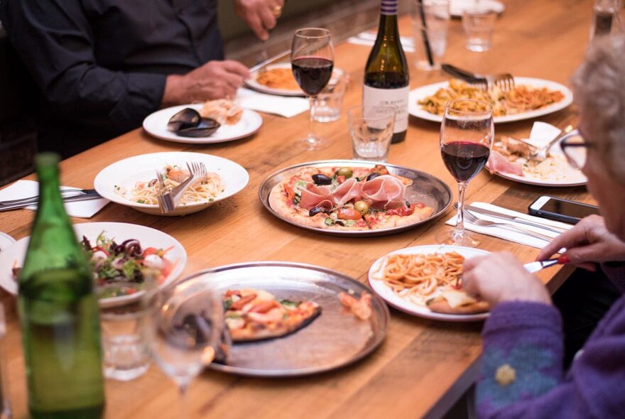 Looking at a table with diners eating pizza, pasta and glasses of red wine.