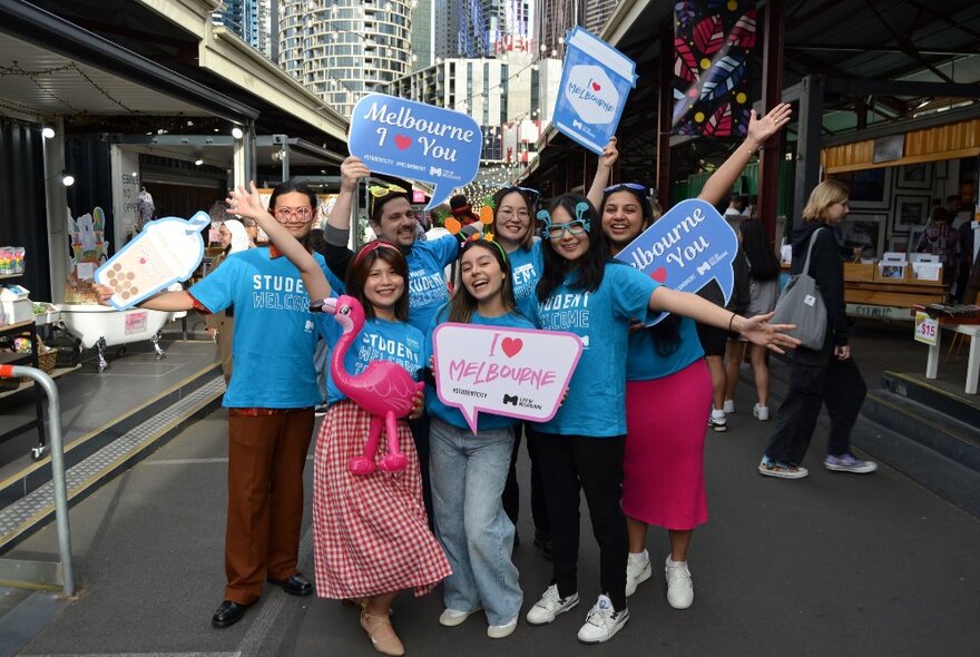 A bunch of young people all wearing the same kind of blue t-shirt, holding signs that say 'I love Melbourne' at the Queen Victoria Market.