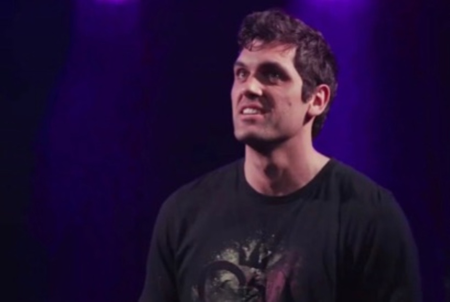 Comedian, Elijah Dries, wearing a black t-shirt in front of a dark purple background.