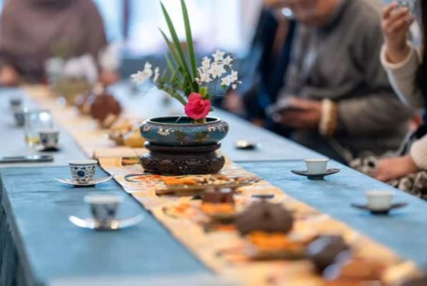 A traditional Chinese tea gathering or ceremony setup with a long table, small tea cups, and a floral centrepiece, with blurred figures in the background.