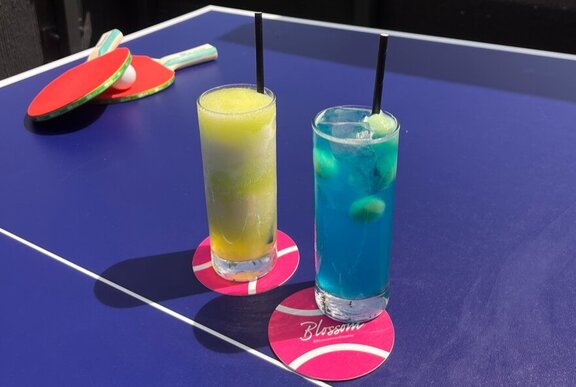 Two cocktails in tall glasses resting on pink tennis ball shaped coasters on a blue ping pong table.