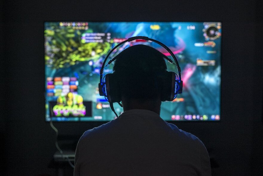 A silhouette of a person wearing headphones in front of a brightly lit computer screen.
