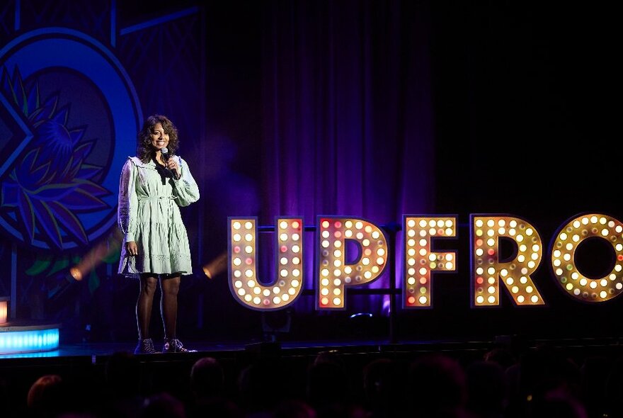 A comedian standing on a stage talking into a microphone, behind her is part of a sign spelling out the letters UPFRO in lights.