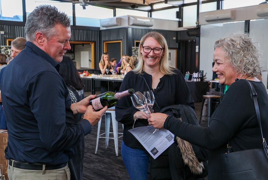 Three people smiling and interacting while the man pours champagne into the women's glasses. 