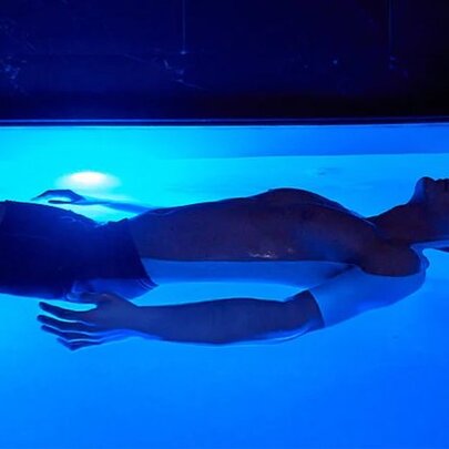 A person experiencing float therapy inside an open float pool or cabin illuminated with blue ambient light.