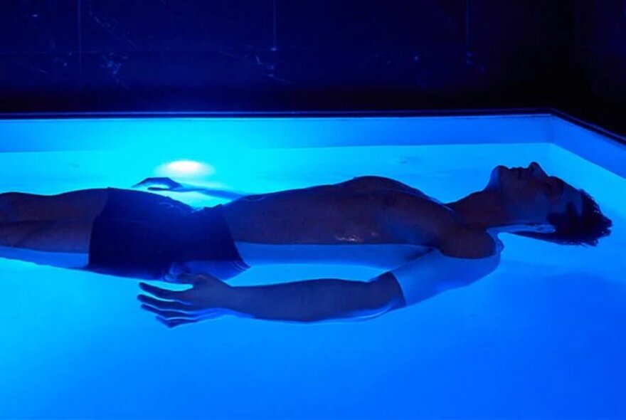 A person experiencing float therapy inside an open float pool or cabin illuminated with blue ambient light.