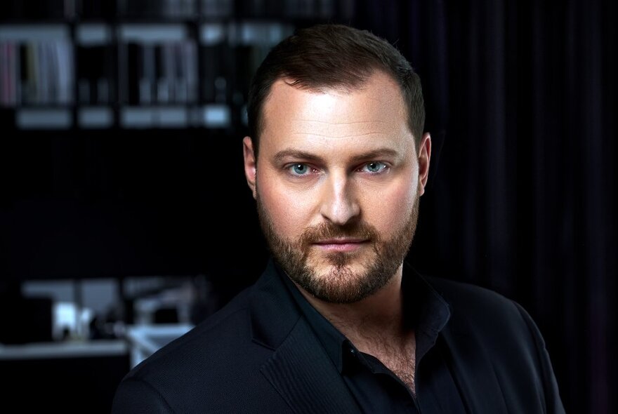 Tenor Michael Petruccelli looking intently straight ahead, wearing a dark suit jacket, with short brown hair, a beard and blue eyes.