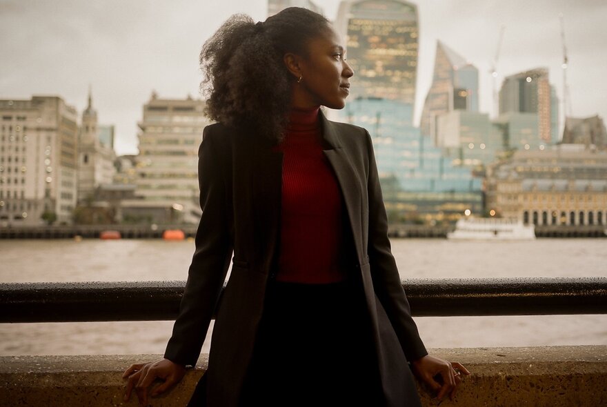 Author Natasha Brown standing in profile beside the Thames with London City buildings in the background.