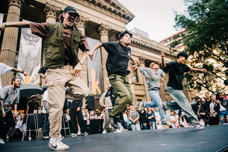 Four street dancers in front of the State Library Victoria, dancing and moving around  on the forecourt area, with a crowd of people watching them perform.