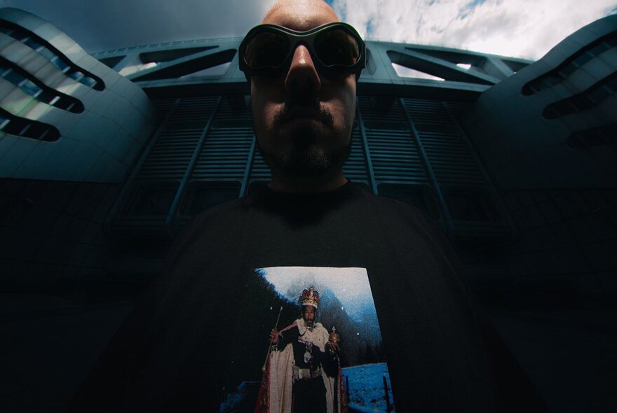 Foreshortened image taken from below of a man in sunglasses, wearing a black t-shirt with an image printed on it, a building looming large behind him.