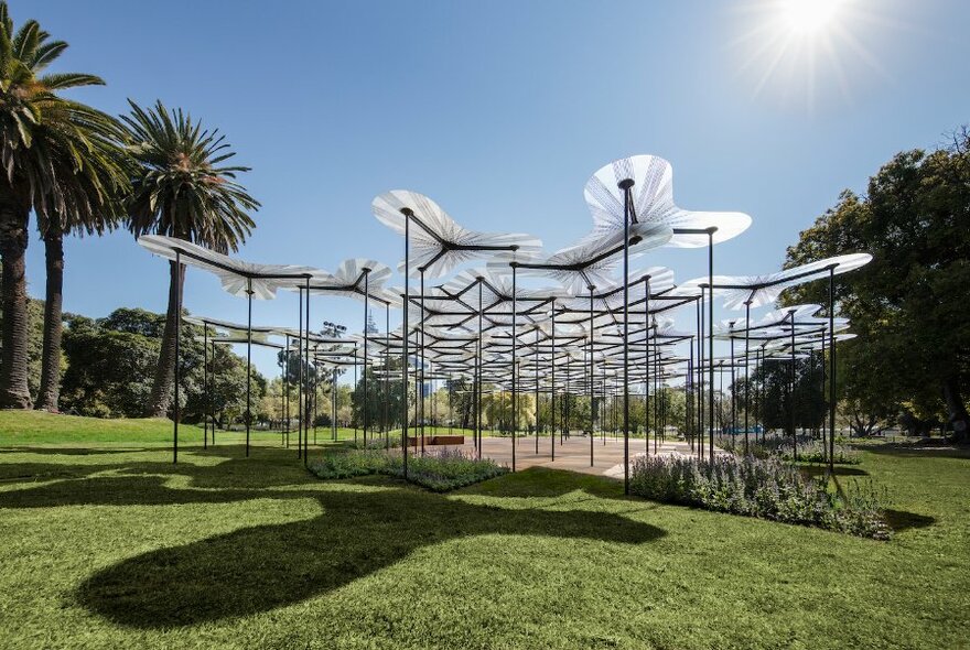 An open structure made up of black poles holding up flat, petal-shaped canopies, surrounded by parkland.