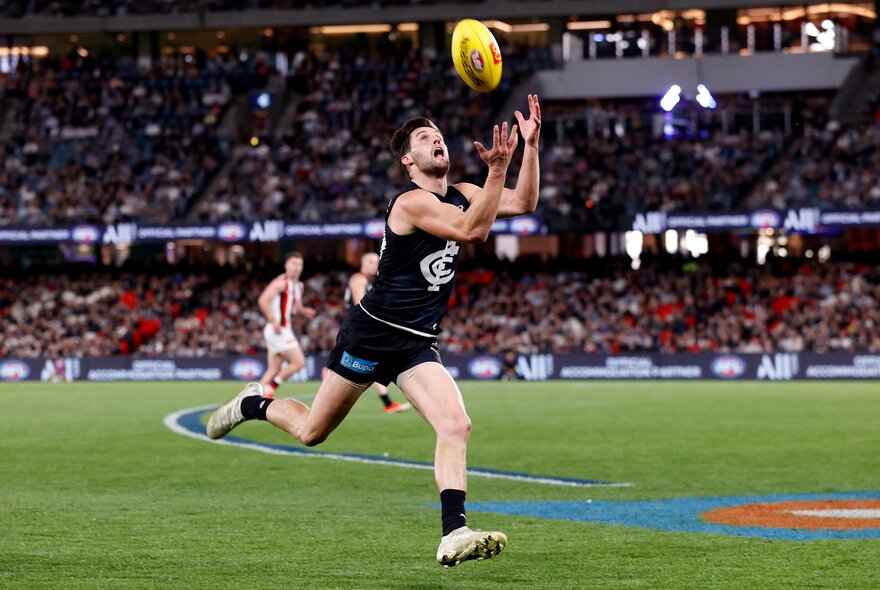 A Carlton AFL player running and looking up, with arms and hands raised, about to mark the footy, with blurred fans in the stadium 