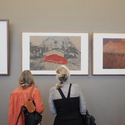 The back of two woman looking at framed art on the wall of a gallery