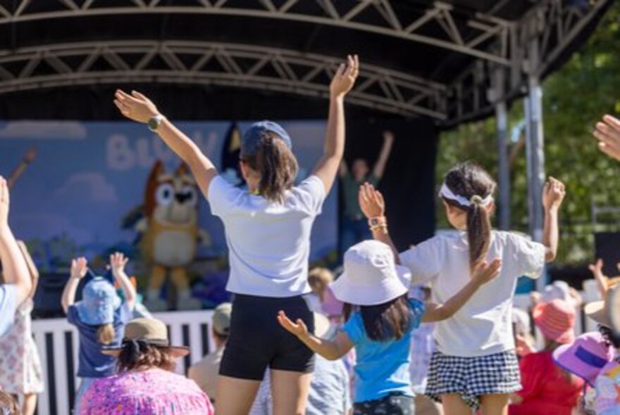 Children waving their arms watching a Bluey stage show outdoors.