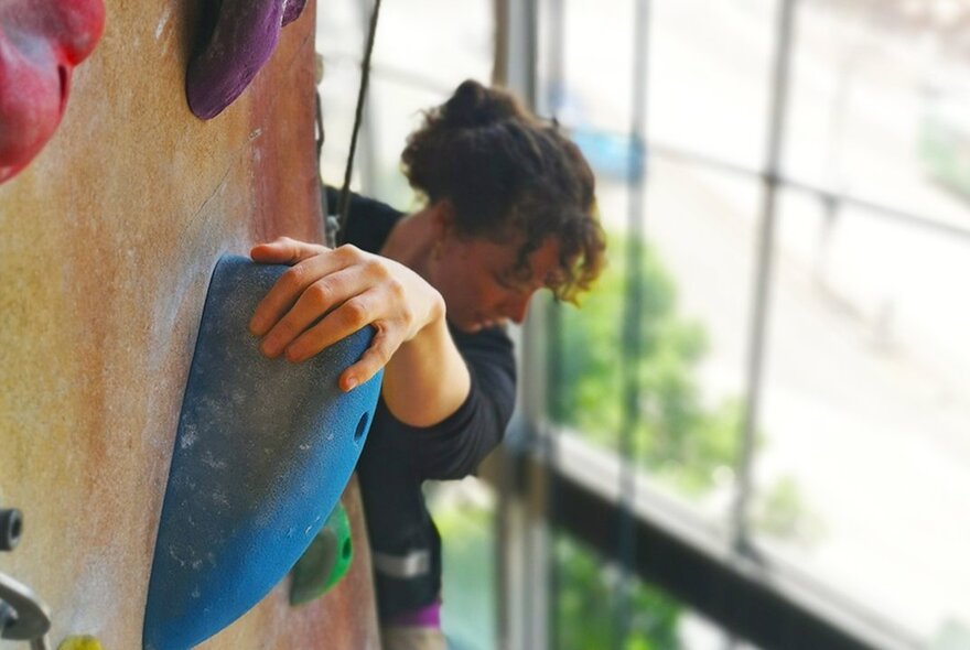 A person indoor climbing, using their hand to hold onto a large grip mounted on the vertical wall.