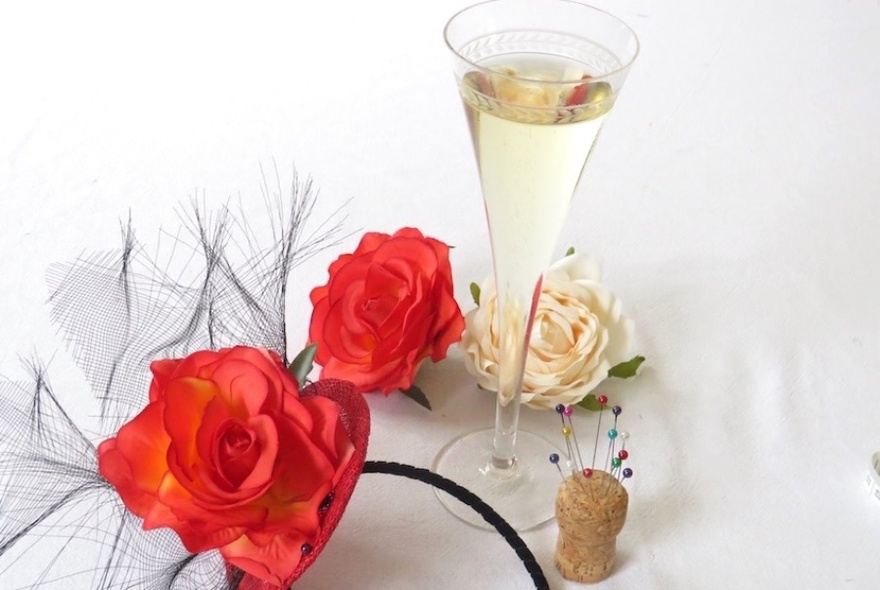 A headpiece featuring red roses and feathers with a white rose, cork with pins and champagne flute beside it. 