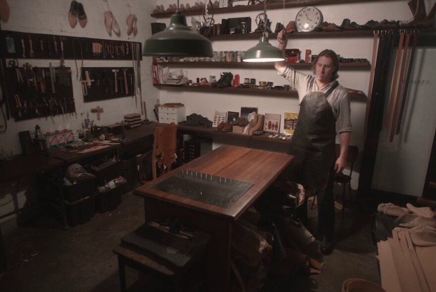A leather workroom with a man in an apron standing behind a workbench, shelves behind him filled with tools and leather pieces.