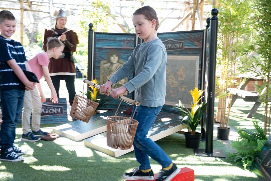 Kids doing a challenge with a girl balancing rocks in two baskets attached to a rod. 