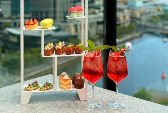 Display of cakes with two glasses of red drink, Yarra River in background.