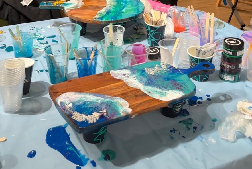 A crafts table with pots of resin and paint, with a wooden board decorated with blue resin.