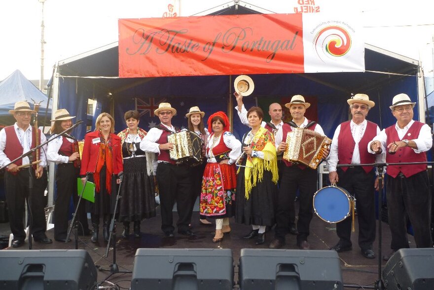 Performers dressed in traditional Portuguese outfits standing on an outside stage with banner.
