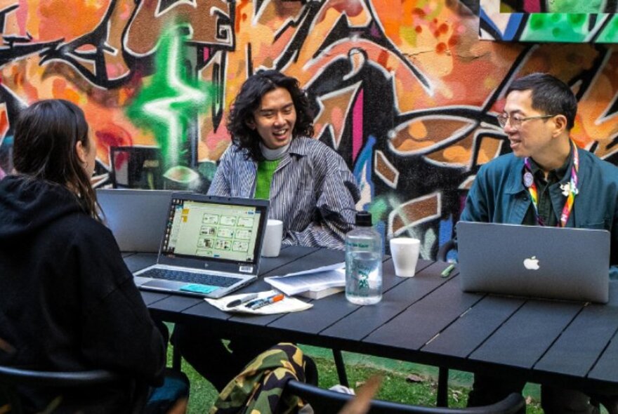 Three people seated at a black outdoor table with laptops in front of them, a graffiti mural on the wall behind them.