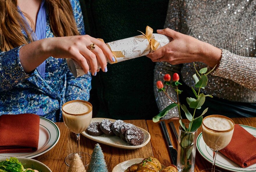 Two hands each pulling the end of a Christmas bon bon cracker, above a table that contains two drinks and a plate of sweet biscuits.