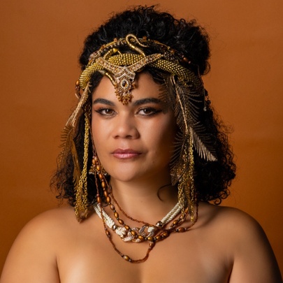 A woman dressed a Cleopatra with bare shoulders and a snake patterned headband against a brown background. 