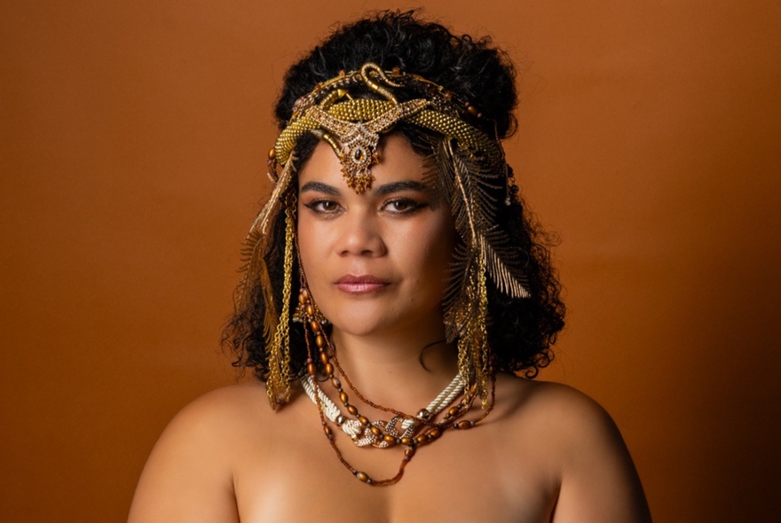 A woman dressed a Cleopatra with bare shoulders and a snake patterned headband against a brown background. 