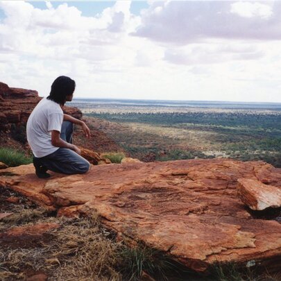 A Japanese man seated on a red rock overlooking a plateau.
