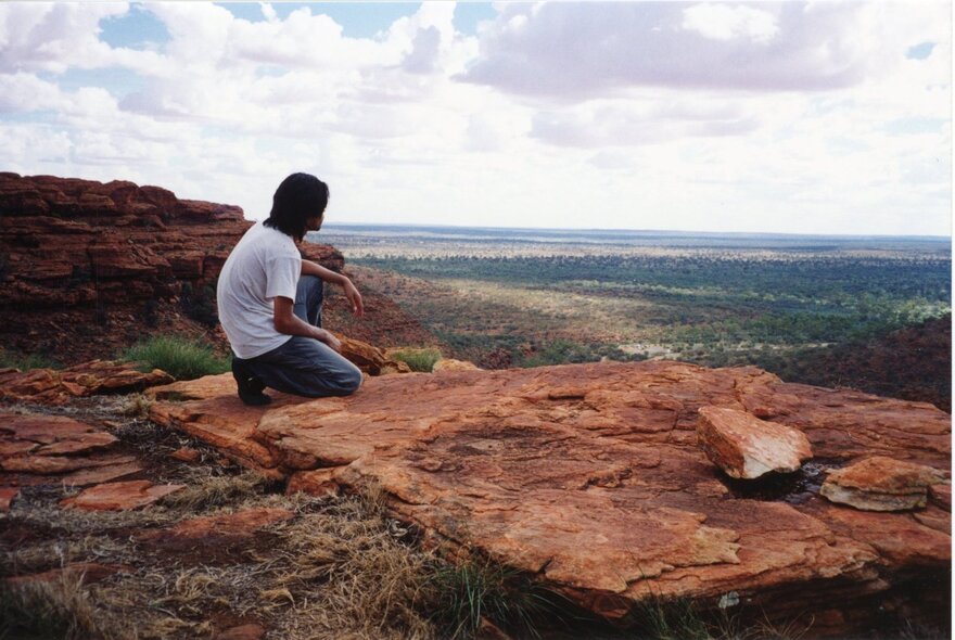 A Japanese man seated on a red rock overlooking a plateau.